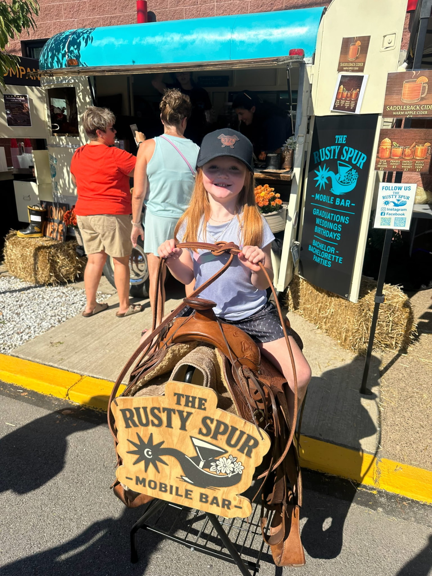 A smiling little girl sitting on the Saddle Photo Setup in front of The Rusty Spur trailer